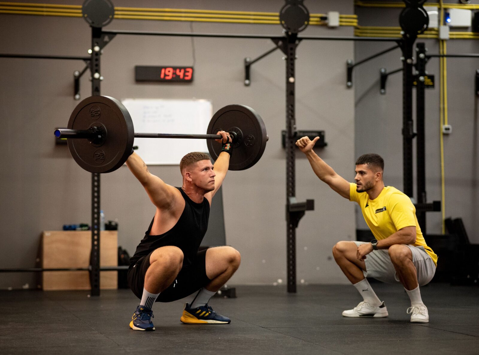 Athlete performing weightlifting exercise under trainer's guidance in a gym setting.